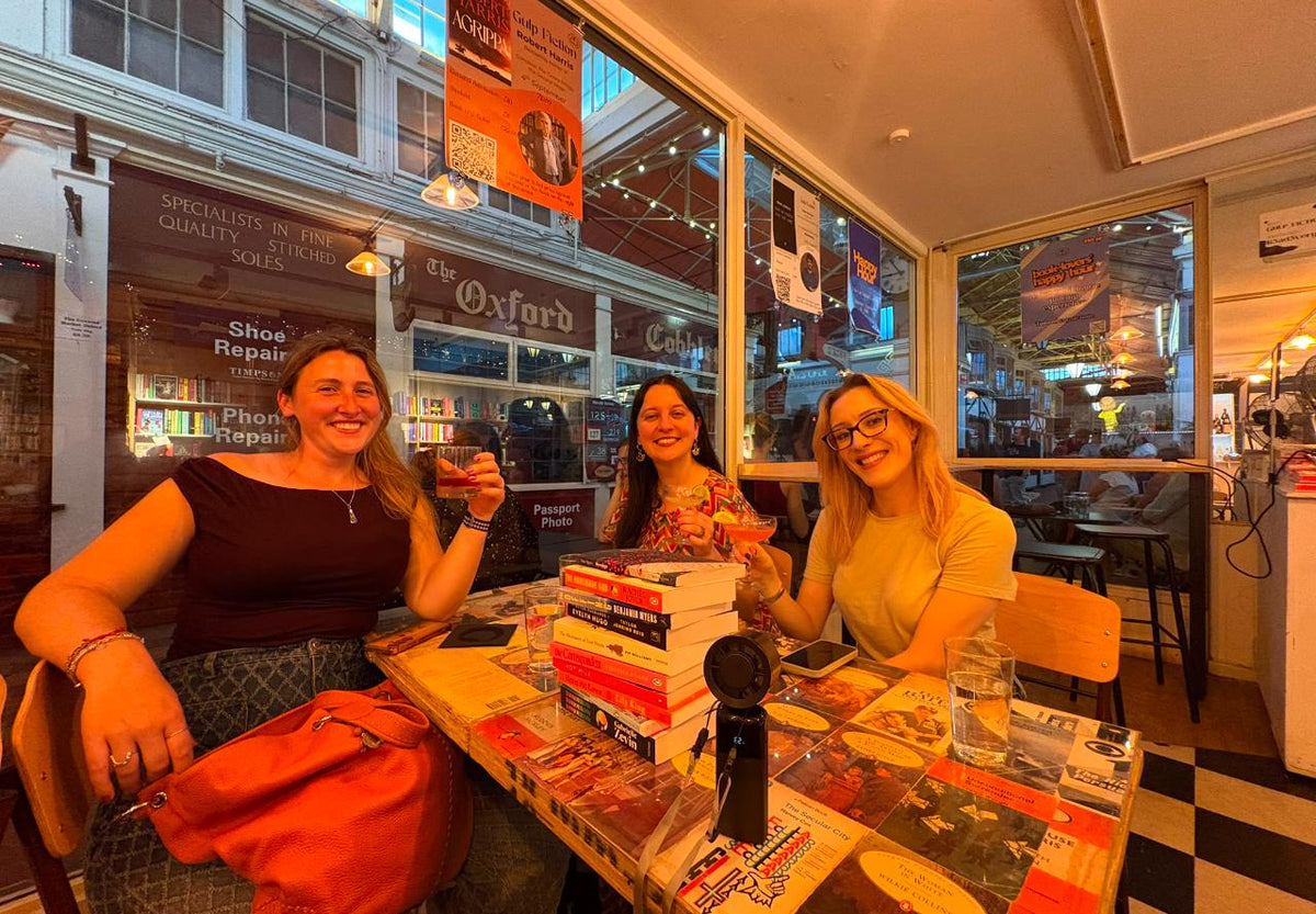 Three customers at the window table in Gulp Fiction, cocktails in hand beside a stack of books, with Oxford's Covered Market visible through the glass